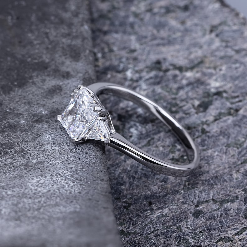 Pode incluir: Um anel de prata com um grande diamante quadrado e dois diamantes triangulares menores de cada lado.