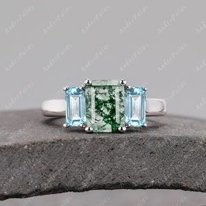 May include: A silver ring featuring a green moss agate centre stone flanked by two light blue stones.