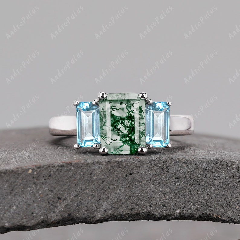 May include: A silver ring featuring a green moss agate centre stone flanked by two light blue stones.