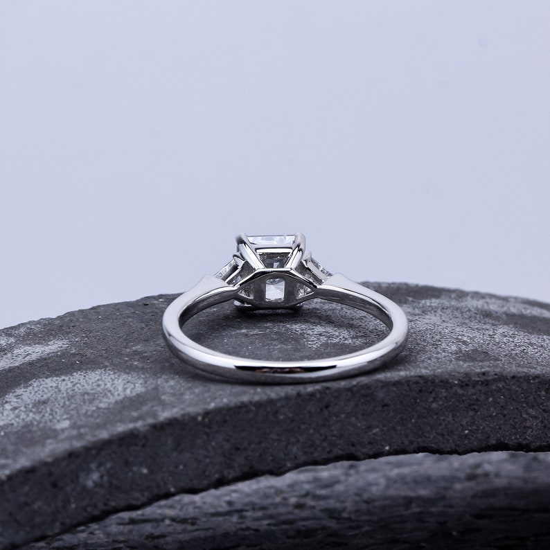 Pode incluir: Um anel de noivado de prata com uma pedra central quadrada e duas pedras menores de cada lado. O anel est&aacute; sobre uma superf&iacute;cie de pedra cinza.