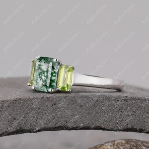 May include: A silver ring featuring a green and white moss agate gemstone, flanked by two smaller green gemstones. The ring is displayed on a grey stone surface.