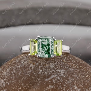 May include: A silver ring featuring a green moss agate emerald cut gemstone, flanked by two smaller, light green peridot gemstones.