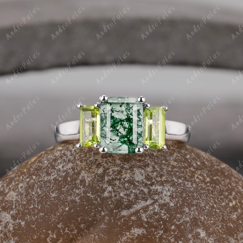 May include: A silver ring featuring a green moss agate emerald cut gemstone, flanked by two smaller, light green peridot gemstones.