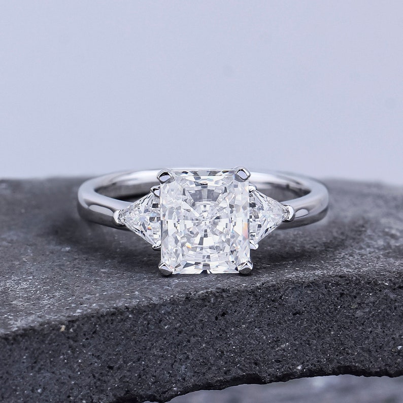 Pode incluir: Um anel de noivado de prata com um grande diamante central quadrado e dois diamantes triangulares menores de cada lado.