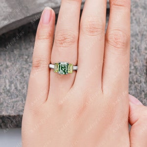 May include: A three-stone engagement ring featuring a large emerald-cut green gemstone, flanked by two smaller square-cut yellow gemstones. The ring is set in a silver band.