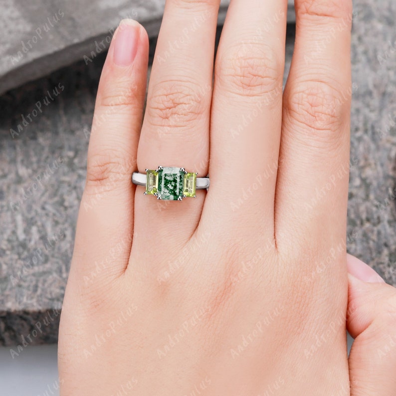 May include: A three-stone engagement ring featuring a large emerald-cut green gemstone, flanked by two smaller square-cut yellow gemstones. The ring is set in a silver band.