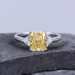 Pode incluir: Um anel de prata com um grande diamante amarelo retangular no centro e dois diamantes triangulares menores de cada lado.