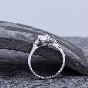 Pode incluir: Um anel de noivado de prata com um grande diamante de corte almofada e dois diamantes menores de cada lado. O anel est&aacute; colocado sobre uma superf&iacute;cie de pedra cinza.