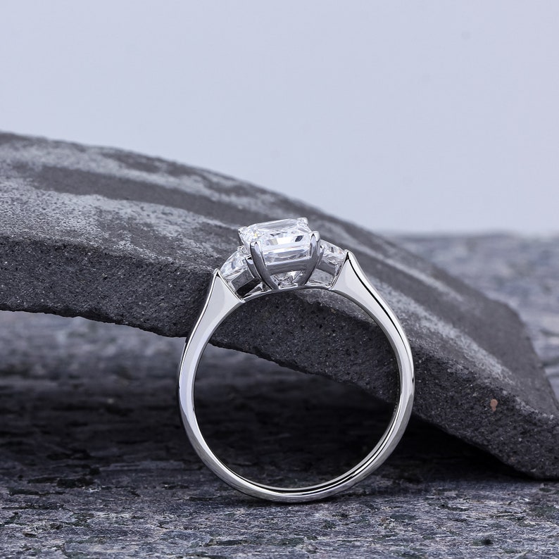 Pode incluir: Um anel de noivado de prata com um grande diamante de corte almofada e dois diamantes menores de cada lado. O anel est&aacute; colocado sobre uma superf&iacute;cie de pedra cinza.
