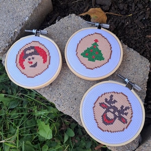 May include: Three small, round, wooden embroidery hoops with white fabric stretched across them. Each hoop features a different Christmas-themed design: a snowman, a Christmas tree, and a reindeer. The designs are stitched in red, green, and brown thread.