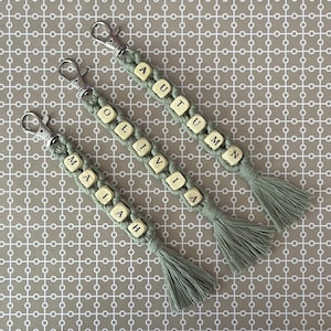 May include: Three sage green macrame keyrings with silver clasps and tassels. Each keyring has a name spelled out with wooden letter beads. The names are: MAIAH, OLIVIA, and AUTUMN.