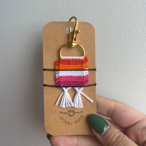 May include: A handmade keychain with a woven design in orange, white, and pink, attached to a gold-coloured clasp and ring. The keychain is secured to a brown card with the text "HOBBY SHOP" and two black strings.