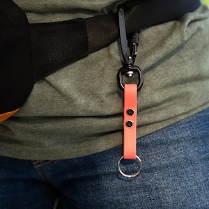 May include: A pink leather key chain with a silver key ring and a black metal clip attached to a black belt.