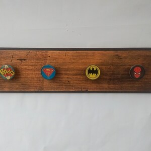 May include: A wooden wall-mounted coat rack with four hooks. Each hook is decorated with a different superhero logo:  "Bang", Superman, Batman, and Spiderman.