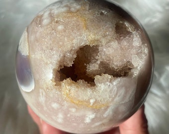 Puede incluir: Una gran esfera de geoda de ágata rosa con una abertura natural que revela un interior forrado de cristales. La geoda tiene un exterior blanco y rosa claro con un toque de morado.