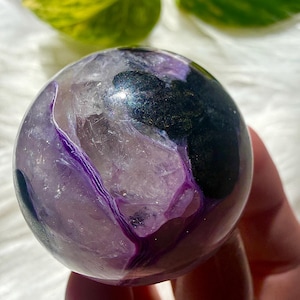May include: A polished spherical stone featuring a translucent light purple amethyst base with deep violet veining and a prominent, opaque dark black inclusion that has a smooth, reflective surface texture across its rounded form.