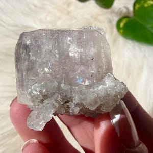 May include: A raw, uncut crystal cluster with a pale lavender hue. The crystal has a rough, textured surface and a crystalline structure. The base of the crystal has a lighter, more opaque appearance. The crystal is held in a hand.