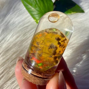 May include: A clear glass vial containing a colourful stone. The stone shows yellow, orange, and red hues with dark brown markings. The vial has a cork base and a small, clear loop at the top. The design is natural and decorative, suitable for display.
