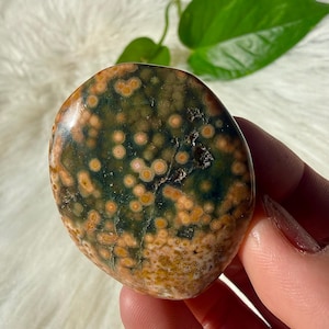 May include: Polished ocean jasper stone with a mix of green, brown, and orange colors. The stone has a rounded shape and a speckled pattern. The surface is smooth and reflective, with a natural, earthy appearance.