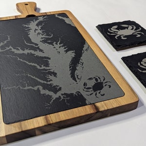 May include: A black slate serving board with a wood frame and a map of the Chesapeake Bay. Two black slate coasters with a crab design are included.
