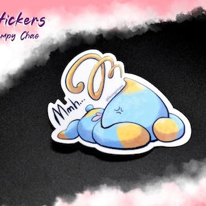 May include: A blue and yellow cartoon Chao character with a grumpy expression. The Chao is lying down with its eyes closed and has a thought bubble above its head that says "Mmm..." The sticker is labeled "Stickers Grumpy Chao".