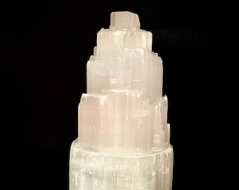 May include: A white selenite crystal tower with a layered, stacked design. The crystal is smooth and polished, with a translucent appearance.