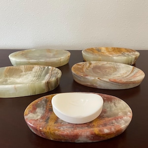 May include: Five oval-shaped onyx soap dishes in shades of green, brown, and red. One dish holds a white soap. The natural stone has unique veining patterns. These decorative dishes are ideal for holding soap or small items.