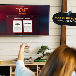 May include: A television screen displays the game Discover Your Kink, featuring a red and black interface with two cards titled Sloppy Blowjob and Spanking. A black speech bubble graphic with yellow and white text reads Play on the big screen.