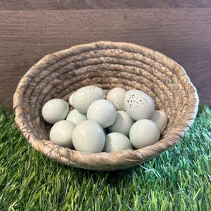 May include: A woven basket filled with speckled, light-green eggs. The eggs are a soft, muted green color with darker speckles. The basket is made of a natural, light-brown woven material. The basket sits on a bed of green artificial grass.