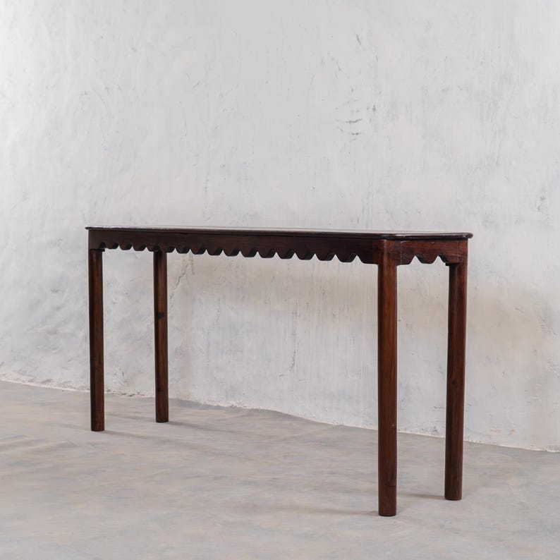 May include: A dark brown, narrow console table with a scalloped edge and four straight legs. The table is made of wood and stands approximately 30 inches tall.