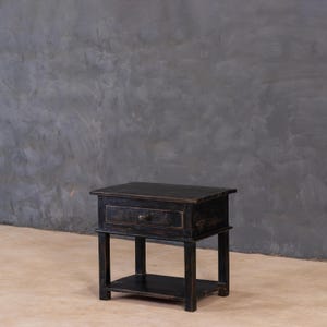 Könnte beinhalten: Ein kleiner, schwarzer Nachttisch aus Holz mit einer Schublade und einem unteren Regal. Der Nachttisch hat eine Used-Optik, die ihm ein rustikales Aussehen verleiht. Die Schublade hat einen runden Metallknopf. Der Nachttisch ist etwa 61 cm hoch.