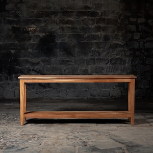 May include: A long, rectangular wooden console table with a lower shelf. The table is made of natural wood with a light brown finish. It is standing on a concrete floor in front of a dark gray brick wall.