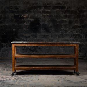 Peut inclure: Une console en bois rustique avec un dessus en pierre gris foncé et deux étagères. La table a une finition marron chaud et repose sur de petites roulettes noires, idéale pour une cuisine ou un salon.