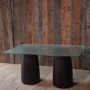 Pode incluir: Uma mesa retangular moderna com um tampo de vidro transparente e dois suportes de madeira castanha escura em forma de barril. A mesa está colocada contra uma parede rústica de tábuas de madeira. A mesa tem cerca de 152 cm de comprimento e 76 cm de largura.