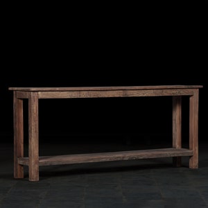 Puede incluir: Una mesa consola de madera rústica con un estante. La mesa tiene un acabado marrón oscuro y está hecha de madera reciclada. Presenta un diseño sencillo con patas rectas y una tapa rectangular.