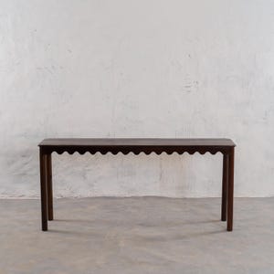 May include: A dark brown wooden console table with a scalloped edge. The table has a rectangular top and four slender legs. The table is approximately 60 inches long, 12 inches wide, and 30 inches tall.