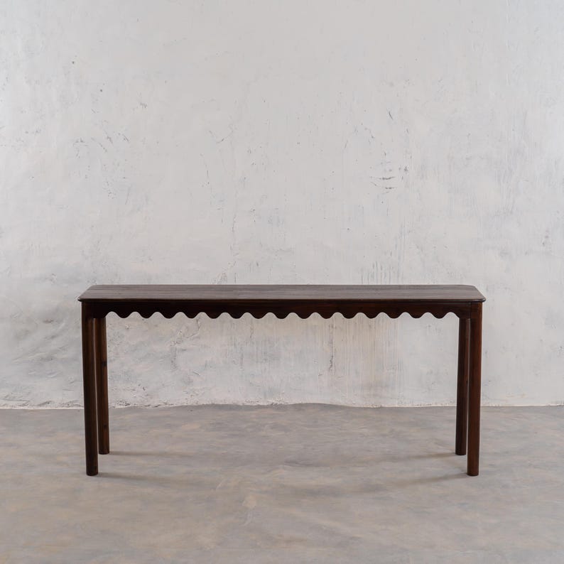 May include: A dark brown wooden console table with a scalloped edge. The table has a rectangular top and four slender legs. The table is approximately 60 inches long, 12 inches wide, and 30 inches tall.
