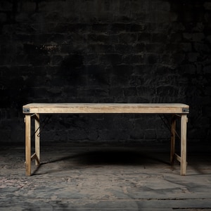 May include: A rustic wooden folding table with black metal hinges. The table is set against a dark brick wall background.