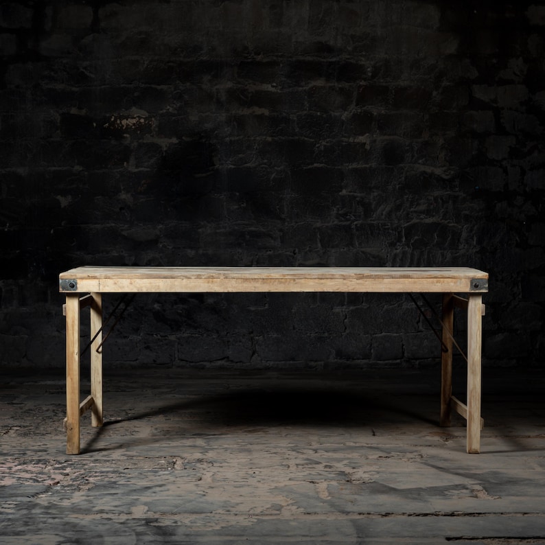 May include: A rustic wooden folding table with black metal hinges. The table is set against a dark brick wall background.