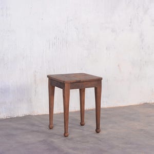 May include: A small, square wooden stool with a dark brown finish. The stool has four legs with rounded feet. The top of the stool is a square shape. The stool is set against a white wall and a gray floor.