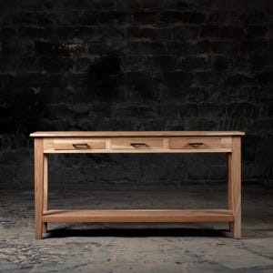 May include: A wooden console table with three drawers and a lower shelf. The table has a natural wood finish and black metal drawer pulls.