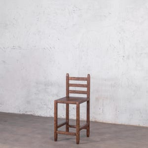 Puede incluir: Un taburete alto de bar de madera con acabado marr&oacute;n oscuro. El taburete tiene un asiento cuadrado, cuatro patas y un respaldo con tres listones horizontales. El dise&ntilde;o es sencillo y r&uacute;stico, adecuado para una cocina o bar.