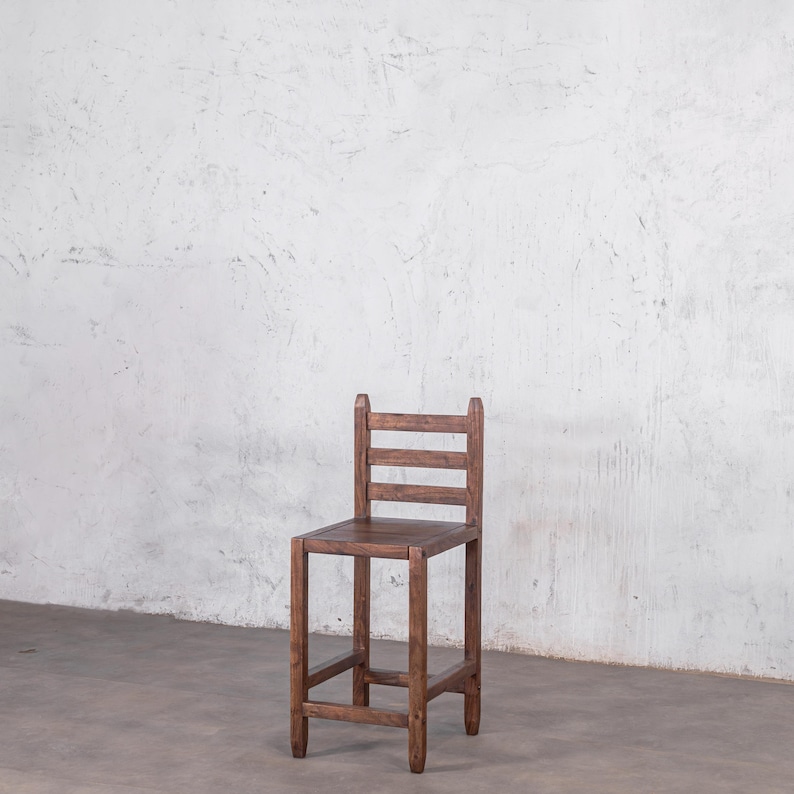 Puede incluir: Un taburete alto de bar de madera con acabado marr&oacute;n oscuro. El taburete tiene un asiento cuadrado, cuatro patas y un respaldo con tres listones horizontales. El dise&ntilde;o es sencillo y r&uacute;stico, adecuado para una cocina o bar.