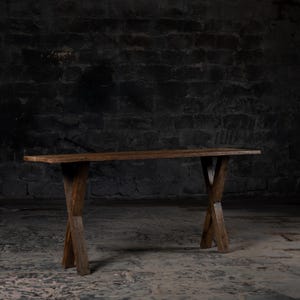 Pode incluir: Uma mesa de madeira rústica com um tampo longo e retangular e uma base simples em forma de X. A mesa é feita de madeira marrom escura e tem um acabamento natural e desgastado.