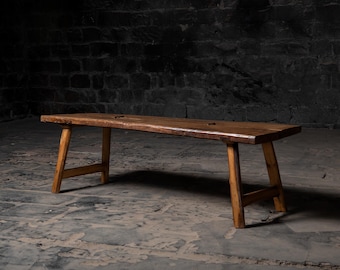 May include: A rustic wooden bench with a long, rectangular top and four simple legs. The bench is made of dark brown wood and has a natural, weathered finish.