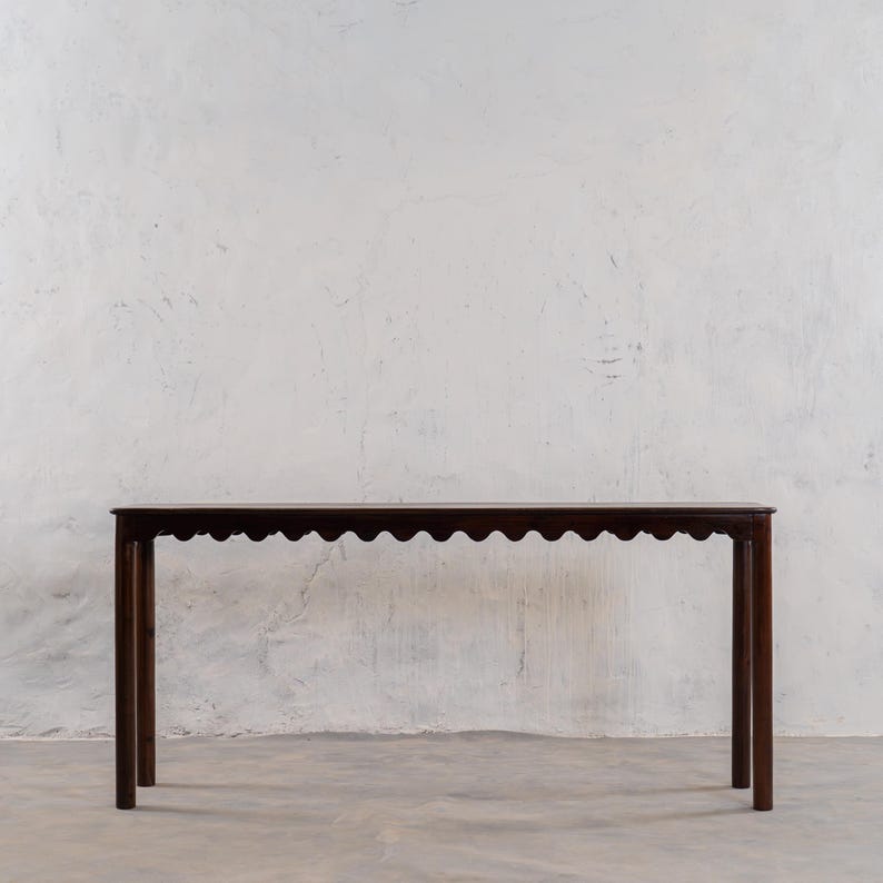May include: A dark brown, rectangular console table with a scalloped edge. The table has four straight legs and is set against a white textured wall. The table is approximately 60 inches long.