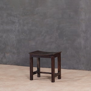 Könnte beinhalten: Ein dunkelbrauner Holzstuhl mit einer geschwungenen Sitzfläche und vier quadratischen Beinen. Der Sitz hat horizontale Lamellen. Der Hocker ist aus dunklem Holz gefertigt und hat ein schlichtes, rustikales Design. Der Hocker steht auf einer hellbraunen Oberfläche mit einer grauen Wand im Hintergrund.
