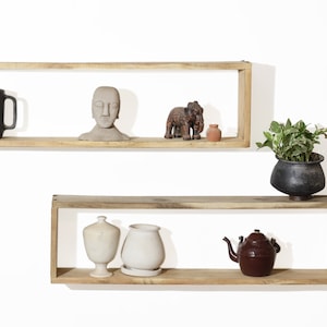 Peut inclure: Deux étagères en bois avec un cadre rectangulaire, montées sur un mur blanc. L'étagère supérieure a une tasse noire, un buste gris, une petite figurine d'éléphant et un petit pot en terre cuite. L'étagère inférieure a un vase blanc, un pot blanc avec une plante et une théière brune.