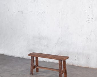 May include: A small, rustic wooden bench with a rectangular seat and four sturdy legs. The bench is made of weathered wood, showcasing natural grain and knots. The design is simple and functional, suitable for indoor or outdoor use.
