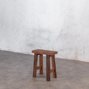 Könnte beinhalten: Ein kleiner, rustikaler Holzschemel mit rechteckiger Sitzfläche und vier stabilen Beinen. Der Hocker ist aus dunkelbraunem Holz gefertigt, das die natürliche Maserung und Textur zeigt. Er steht auf einem grauen Betonboden vor einer weißen Wand.